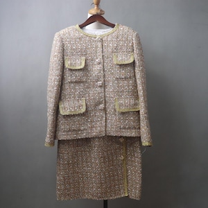 May include: A two-piece tweed suit in beige, brown, and white, with gold trim. The jacket has four pockets and a round neckline. The skirt is knee-length. The suit is displayed on a wooden hanger against a gray background.