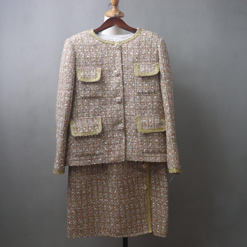 May include: A two-piece tweed suit in beige, brown, and white, with gold trim. The jacket has four pockets and a round neckline. The skirt is knee-length. The suit is displayed on a wooden hanger against a gray background.