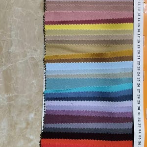 Puede incluir: Una muestra de tela multicolor que muestra una amplia gama de colores, desde beige claro hasta verde azulado intenso y burdeos. La muestra está marcada con números en el lateral, lo que indica diferentes muestras de color. La tela parece ser un material texturizado.