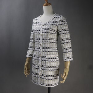 May include: A long-sleeved, button-down dress in a tweed fabric. The dress features a V-neckline and is primarily white with black and tan stripes. It has two front pockets and is displayed on a mannequin.