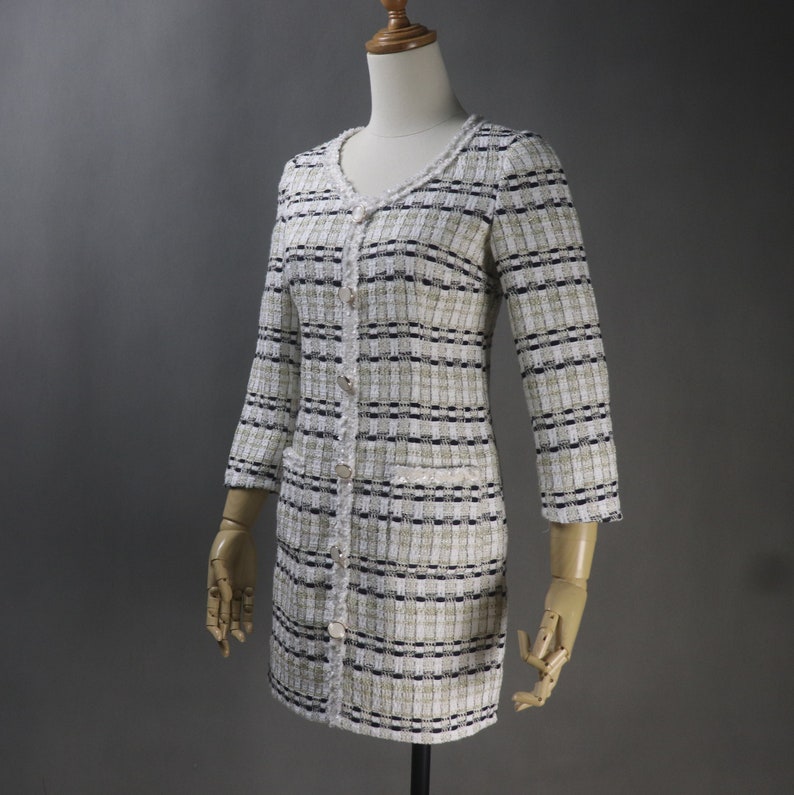 May include: A long-sleeved, button-down dress in a tweed fabric. The dress features a V-neckline and is primarily white with black and tan stripes. It has two front pockets and is displayed on a mannequin.