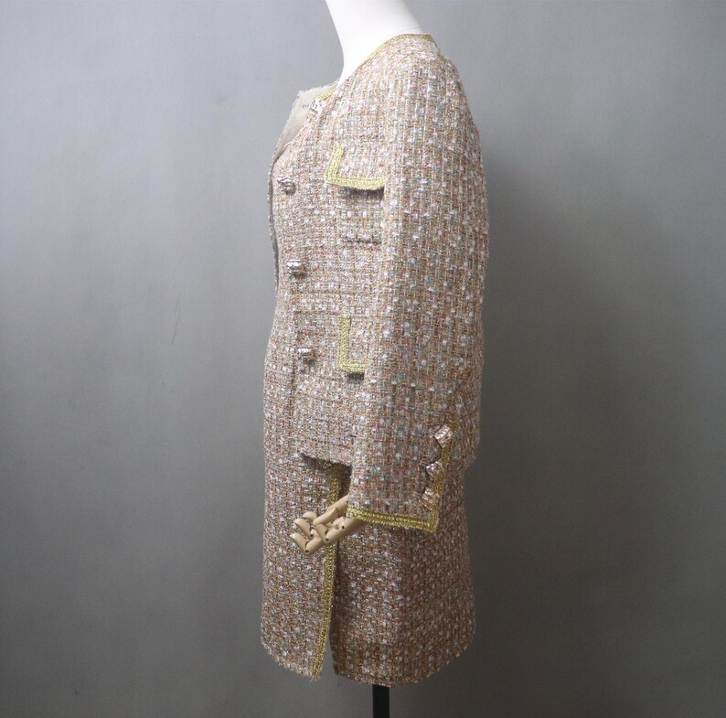 May include: A beige tweed skirt suit with gold trim and buttons. The jacket has pockets and a round neckline. The skirt is knee-length. The suit is displayed on a mannequin.
