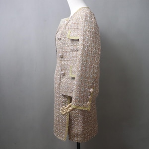 May include: A beige tweed skirt suit with gold trim and buttons. The jacket has a round neckline, pockets, and long sleeves. The skirt is knee-length. The suit is displayed on a mannequin.