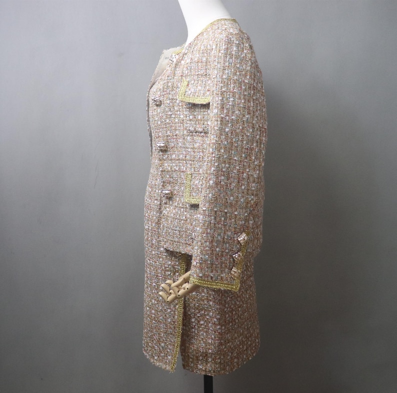 May include: A beige tweed skirt suit with gold trim and buttons. The jacket has a round neckline, pockets, and long sleeves. The skirt is knee-length. The suit is displayed on a mannequin.