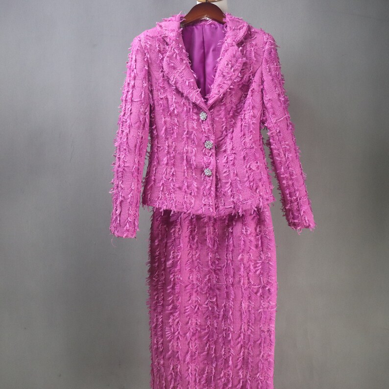May include: A pink tweed skirt suit with a blazer and a skirt. The blazer has a notched collar, long sleeves, and a button closure. The skirt is a pencil skirt with a high waist.