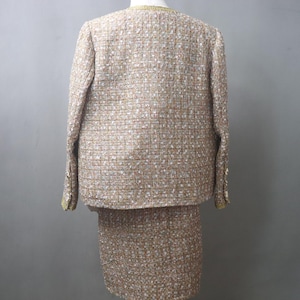 May include: A beige tweed skirt suit. The jacket has long sleeves and gold trim around the collar and cuffs. The skirt is knee-length. The fabric has flecks of white, brown, and red.