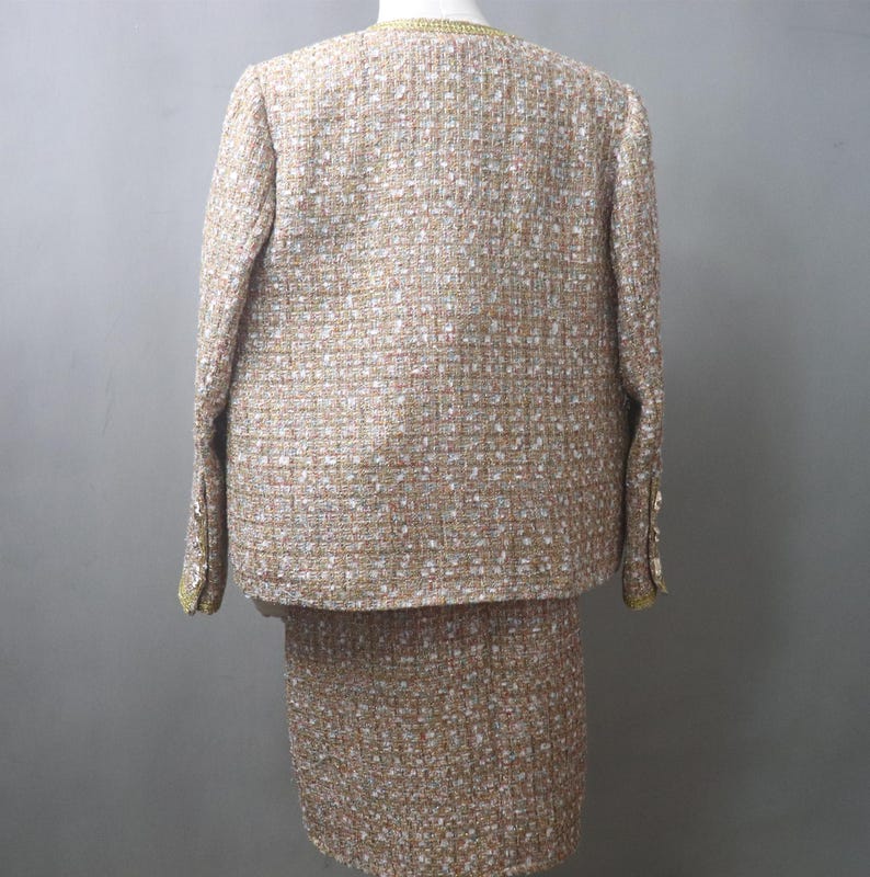 May include: A beige tweed skirt suit. The jacket has long sleeves and gold trim around the collar and cuffs. The skirt is knee-length. The fabric has flecks of white, brown, and red.