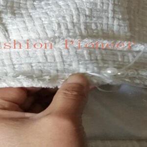 May include: Close-up of a white textured fabric with the text "Fashion Pioneer 15" in orange. The fabric is embellished with small, white pearls along the edge. A hand is visible, touching the fabric.