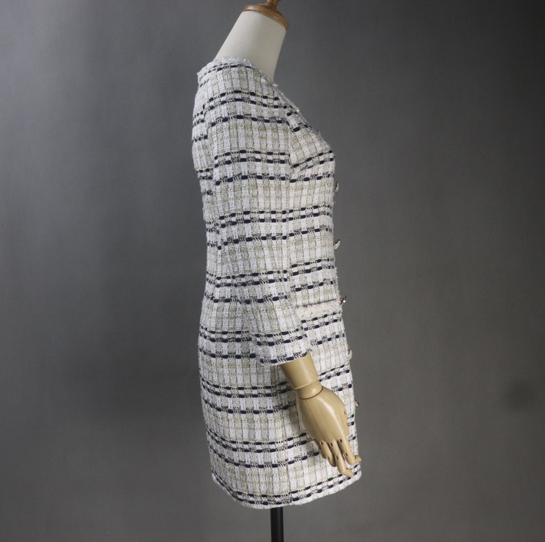 May include: A tweed dress in a cream, black, and gold plaid pattern. The dress features a V-neck, three-quarter sleeves, and decorative buttons down the front. The dress is displayed on a mannequin.