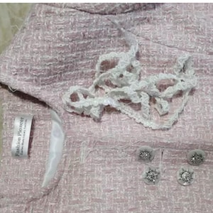 May include: Close-up of a light pink tweed jacket with white lace trim and decorative buttons. The jacket has a label that reads "Fashion Pioneer". The buttons are silver with a floral design.