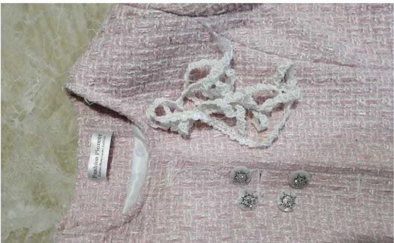 May include: Close-up of a light pink tweed jacket with white lace trim and decorative buttons. The jacket has a label that reads "Fashion Pioneer". The buttons are silver with a floral design.