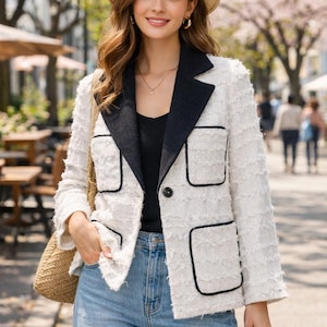 Puede incluir: Una mujer con una chaqueta de tweed blanca con ribetes y bolsillos negros, combinada con vaqueros azules y un sombrero de paja. La chaqueta tiene un cuello negro y un cierre de un solo botón. Un bolso de paja está sobre su hombro.