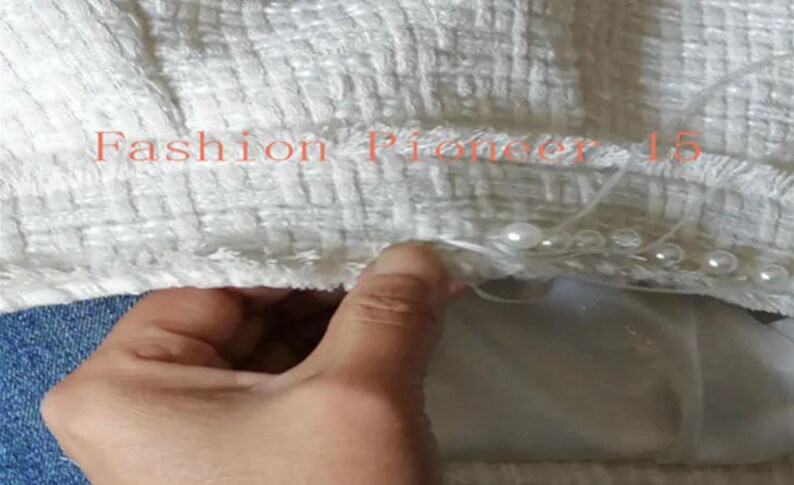 May include: Close-up of a white textured fabric with the text "Fashion Pioneer 15" in orange. The fabric is embellished with small, round, white pearls. A hand is visible, touching the fabric.