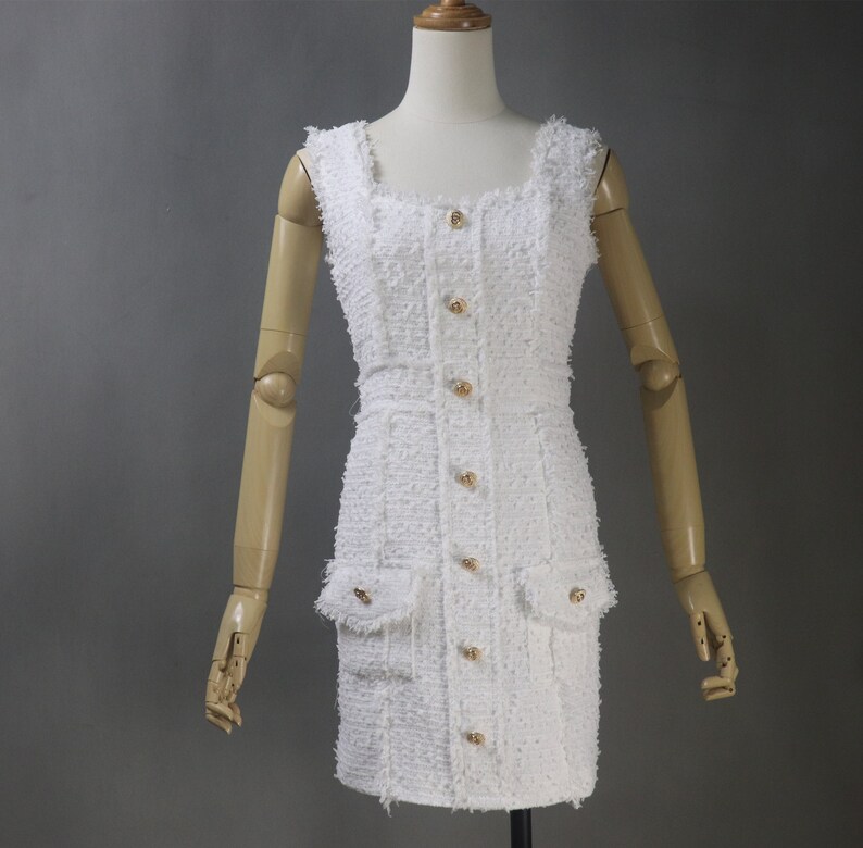 Op de afbeelding: Witte, mouwloze jurk met een vierkante halslijn en een knoopsluiting aan de voorkant. De jurk heeft een tweed-textuur en gouden knopen.