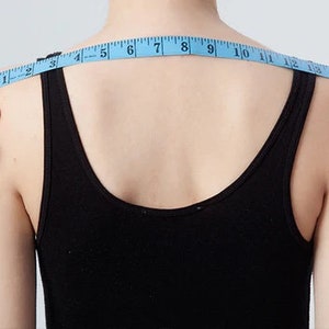 May include: A person is measuring their shoulder width with a blue tape measure. The tape measure reads from 1 to 18 inches.