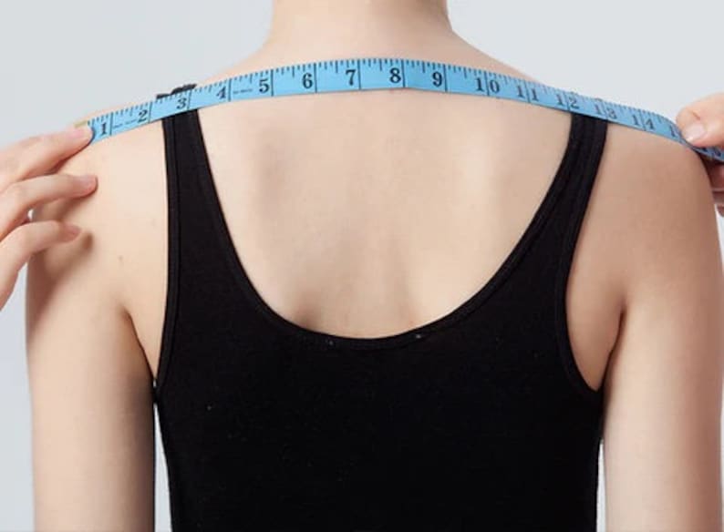 May include: A person is measuring their shoulder width with a blue tape measure. The tape measure reads from 1 to 18 inches.