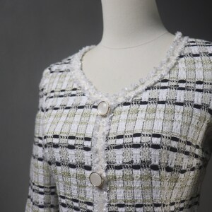 May include: A white, black, and gold tweed jacket with a V-neckline and two large, round, pearlescent buttons. The jacket has a textured weave and a delicate, fringed trim around the neckline and button placket.