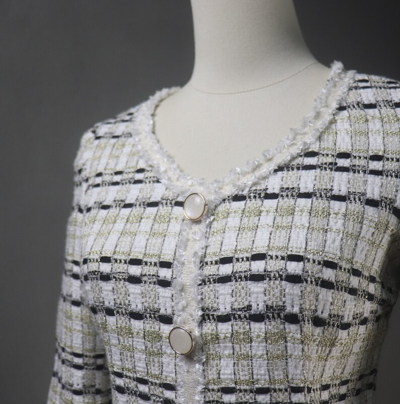 May include: A white, black, and gold tweed jacket with a V-neckline and two large, round, pearlescent buttons. The jacket has a textured weave and a delicate, fringed trim around the neckline and button placket.