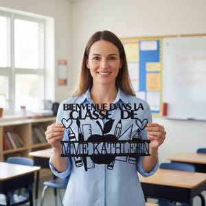 Peut inclure: Une enseigne en métal noir avec le texte "BIENVENUE DANS LA CLASSE DE MME KATHLEEN" et des illustrations sur le thème de l'école. L'enseigne représente une pomme, des crayons, des pinceaux et des cœurs. L'enseigne est tenue par une femme souriante dans une salle de classe.