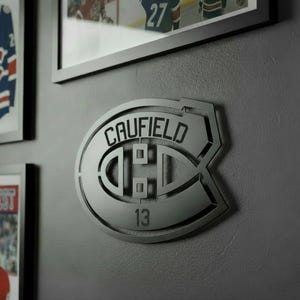 May include: A silver metal wall decoration featuring the Montreal Canadiens logo, with the name "CAUFIELD" and the number "13". The logo is mounted on a grey wall, with framed pictures visible in the background.