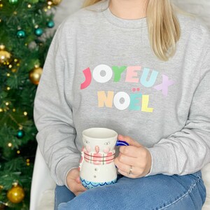 以下が含まれることがあります： 「JOYEUX NOËL」の文字がカラフルにプリントされたグレーのスウェットシャツ。青い持ち手の雪だるまをテーマにしたマグカップを持っています。背景にはオーナメントの付いたクリスマスツリーがあります。