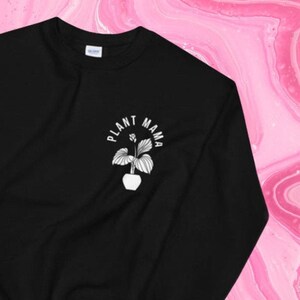 Puede incluir: Sudadera negra con la frase "PLANT MAMA" y una ilustración blanca de una planta en maceta. Esta sudadera de cuello redondo está hecha de un material suave y es adecuada para el uso diario.