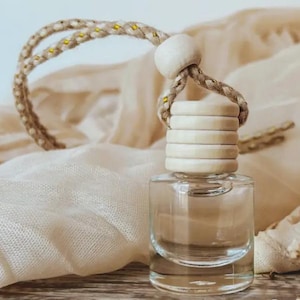 May include: A clear glass bottle with a wooden cap and a braided rope loop. The bottle is filled with a clear liquid and sits on a light-colored surface. The rope loop has gold accents.