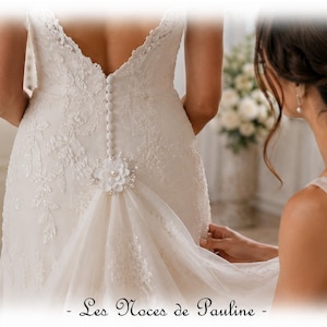 May include: An ivory wedding dress featuring a deep scoop back, a vertical row of fabric-covered buttons, and intricate lace floral embroidery. A decorative floral applique with a central pearl accent sits at the waist, gathering tulle fabric.