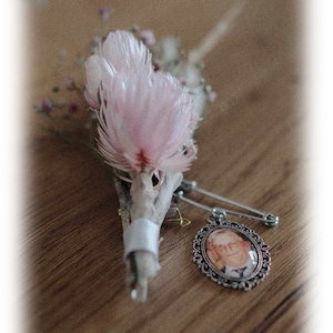 Puede incluir: Un pequeño ramo de flores secas con una flor rosa en el centro, atado con una cinta blanca. Un medallón plateado con una foto de una persona en su interior está sujeto a un imperdible.