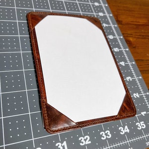 May include: A rectangular brown leather document holder with a white paper insert. The corners of the holder are reinforced with triangular leather pieces. The holder is on a gray cutting mat with measurement markings.