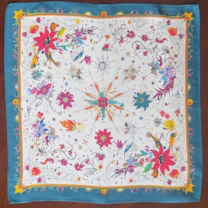 May include: Square silk scarf with a white background and a colorful floral design. The scarf features a teal border and a detailed pattern of flowers, leaves, and abstract shapes in various colors, including red, pink, and yellow.