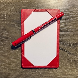 Könnte beinhalten: Ein roter Leder-Notizblockhalter mit einem weißen Notizblock und einem roten Stift. Der Stift ist über dem Notizblock angewinkelt. Der Notizblockhalter hat genähte Kanten und dreieckige Eckakzente.