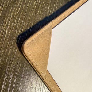 Może przedstawiać: Zbliżenie na brązowy skórzany przedmiot z szytą obwódką. Przedmiot ma zaokrąglony róg i kontrastującą białą powierzchnię. Skóra wydaje się być zamszem lub podobnym miękkim materiałem. Tło to ciemne usłojenie drewna.