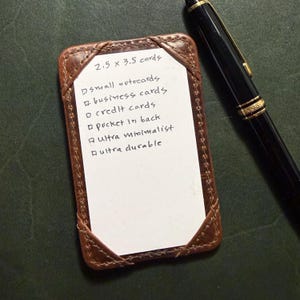May include: A brown leather card holder with a white insert, holding a list of card types. The text reads "2.5 x 3.5 cards". A black pen with gold accents rests beside the holder. The holder is designed for small notecards, business cards, and credit cards.