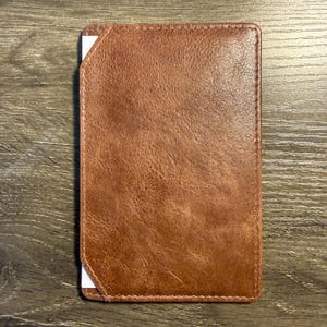 May include: A rectangular brown leather wallet with a textured surface. The wallet has white corner accents and is stitched around the edges. The wallet is lying on a wooden surface.