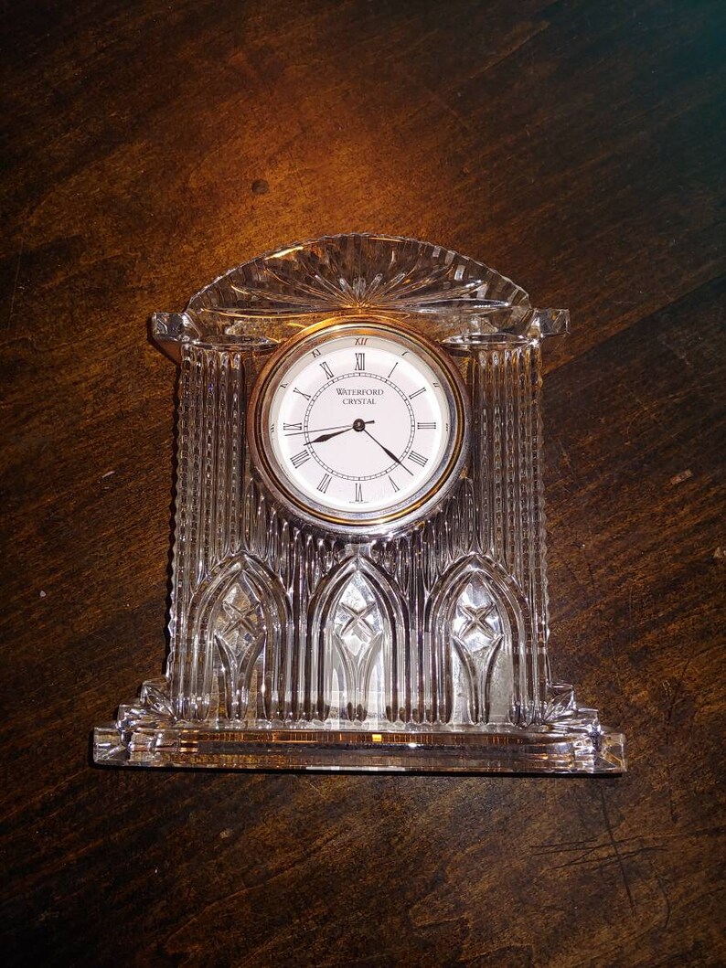 May include: A Waterford Crystal clock with a gold-rimmed face and Roman numeral markings. The clock is made of clear crystal and has a decorative design.