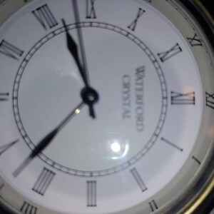 May include: A white clock face with black Roman numeral hour markers and black hands. The clock face has the words "Waterford Crystal" printed on it.
