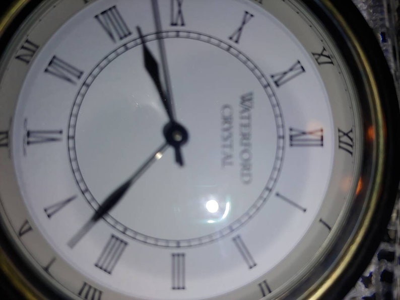 May include: A white clock face with black Roman numeral hour markers and black hands. The clock face has the words "Waterford Crystal" printed on it.
