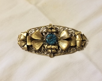 May include: A gold filigree brooch with a blue rhinestone in the center. The brooch is shaped like a bow and has a delicate, intricate design.