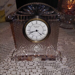 May include: A Waterford Crystal clock with a clear crystal base and a gold-toned face. The clock face has Roman numerals and the words "Waterford Crystal" printed on it.