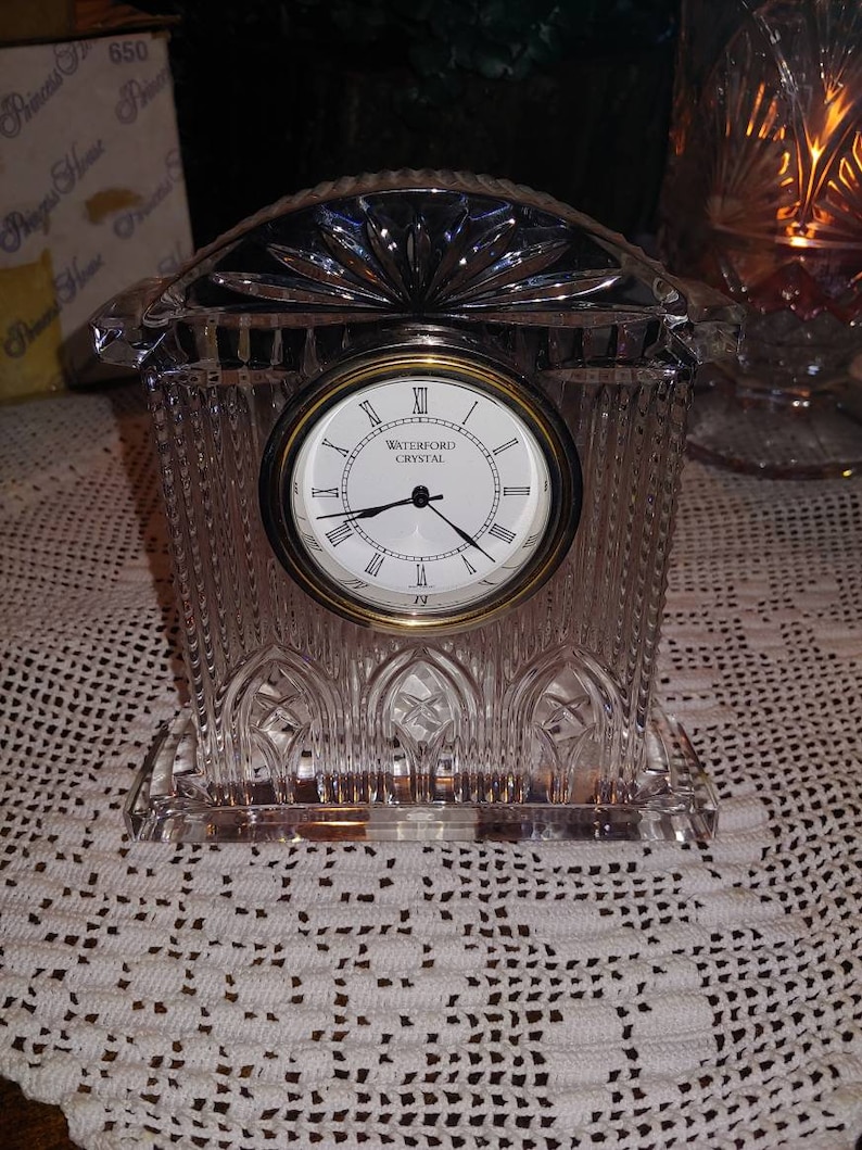May include: A Waterford Crystal clock with a clear crystal base and a gold-toned face. The clock face has Roman numerals and the words "Waterford Crystal" printed on it.