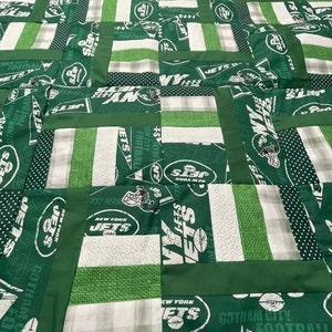 Puede incluir: Una colcha de retazos verde y blanca con el logotipo de los New York Jets repetido en toda la superficie. La colcha está hecha de varias telas, incluyendo cuadros, lunares y verde liso.