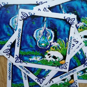 May include: A stack of stickers featuring a blue and green watercolour illustration of a skull with a glowing blue flame above it. The skull is surrounded by green grass and flowers. The stickers have a white border with a decorative blue and green pattern.