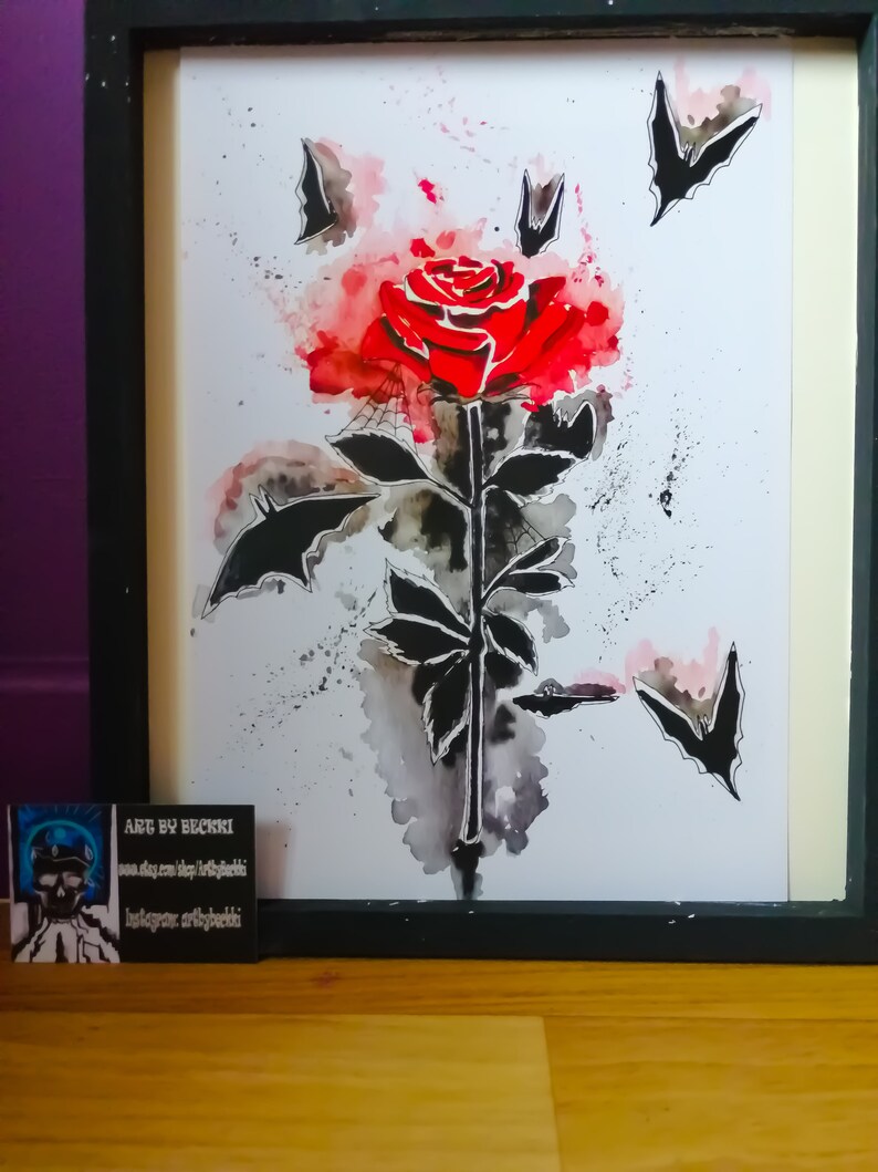 Puede incluir: Una pintura de acuarela de una rosa roja con murci&eacute;lagos negros volando a su alrededor. La rosa est&aacute; pintada en un estilo realista, con un tallo y hojas detalladas. Los murci&eacute;lagos son estilizados y tienen un toque caprichoso. La pintura est&aacute; sobre un fondo blanco.