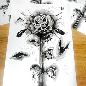 May include: A black and white ink drawing of a rose with a spider web, bats, and beads on the stem. The rose is drawn in a gothic style.