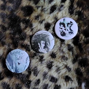 May include: Three round white button pins with black and white illustrations. The first pin features a black and white illustration of a woman with long black hair. The second pin features a black and white illustration of a cemetery with the text "Cemetery Bats". The third pin features a black and white illustration of two figures standing in front of a cross.