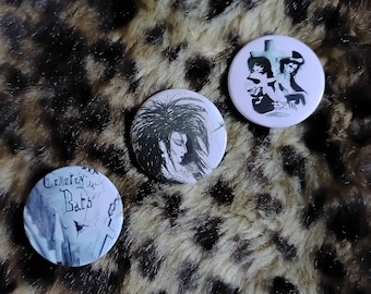 Puede incluir: Tres pines redondos blancos con ilustraciones en blanco y negro. El primer pin presenta una ilustración en blanco y negro de una mujer con cabello negro largo. El segundo pin presenta una ilustración en blanco y negro de un cementerio con el texto "Cemetery Bats". El tercer pin presenta una ilustración en blanco y negro de dos figuras de pie frente a una cruz.