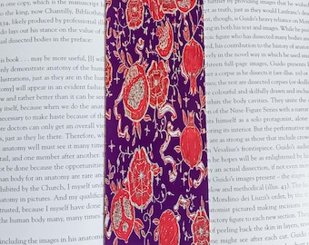 Puede incluir: Un marcapáginas rectangular con un patrón repetido de ilustraciones de granadas rojas sobre un fondo morado oscuro. El marcapáginas está colocado sobre un libro abierto, con texto visible.