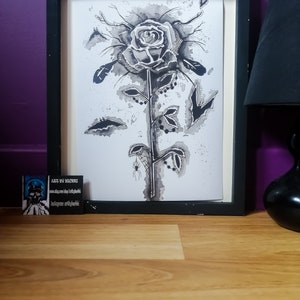 May include: Black and white ink drawing of a rose with a spider web and spider on the stem. The drawing is framed in a black frame and is on a wooden surface.