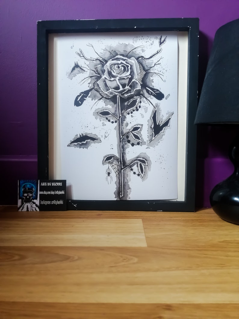 May include: Black and white ink drawing of a rose with a spider web and spider on the stem. The drawing is framed in a black frame and is on a wooden surface.